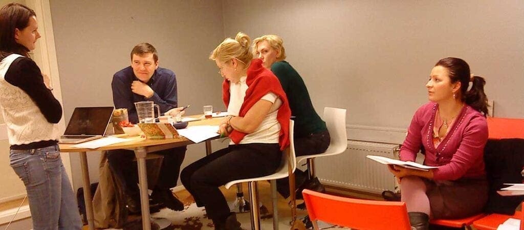 lärare som står och fyra vuxna elever som sitter vid höga bord